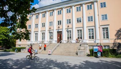 Suureen vaaleankeltaiseen rakennukseen johtaa leveät portaat. Rakennuksen edellä kulkeen ihmisiä. Sää on aurinkoinen.