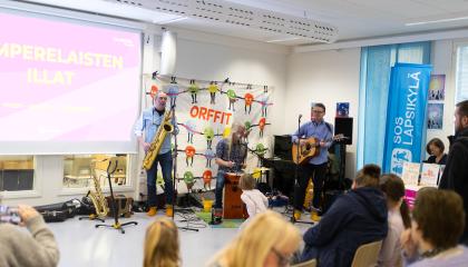 Lastenmusiikkiyhtye soittaa koulun tiloissa. Yleisössä on eri-ikäisiä ihmisiä.