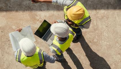 Kolme rakennusalalla työskentelevää henkilöä seisoo pihalla. Heidät on kuvattu ylhäältä päin. Päässä kypärät ja päällä huomioliivit. Yksi osoittaa kädellään eteenpäin, toisella on sylissä läppäri ja kolmannella papereita.