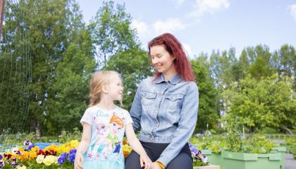 Äiti ja lapsi katsovat toisiaan silmiin ja hymyilevät. Heidän takanaan näkyy lehtimetsää sekä istutuslaatikkoja, joissa on muun muassa värikkäitä orvokkeja.