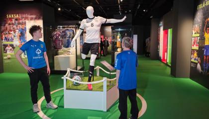 Kaksi lasta katselee mallinukkea, jonka päällä on jalkapalloasu näyttelytilassa.