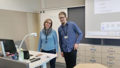 Kaksi henkilöä seisoo luokassa opettajanpöydän luona.