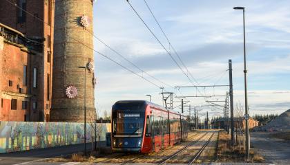 Kuvassa keskellä ratikka, vasemmalla piippu jossa kukiksta muodostuva teos.