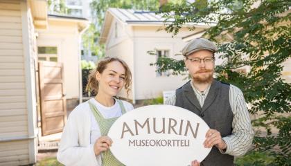 Kaksi vanhanaikaisiin asuihin pukeutunutta henkilöä pitelee käsissään Amurin museokortteli -kylttiä.
