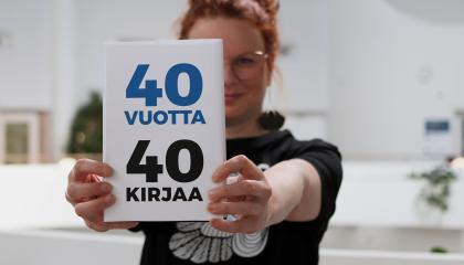 Tuija seisoo Metson parvikerroksessa ja pitää osittain kasvojensa edessä kirjaa, jonka kannessa on teksti 40 vuotta, 40 kirjaa. 