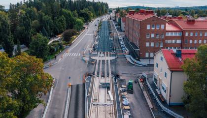 Kuvassa keskellä raitiotietyömaa ja autokaistoja, vihreää vasemmalla ja punatiilinen koulu oikealla. 