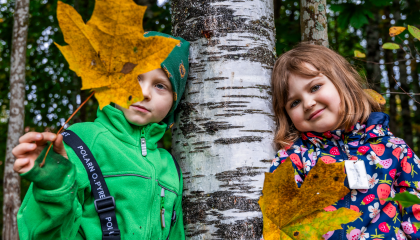 Kaksi lasta nojaa koivuun ja toinen pitää edessään isoa vaahterapuun lehteä.