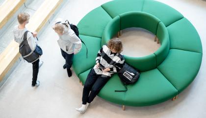 Ylhäältä otettu kuva pyöreästä vihreästä sohvasta, jolla istuu yksi opiskelija. Hänen lähellään seisoo kaksi muuta opiskelijaa.