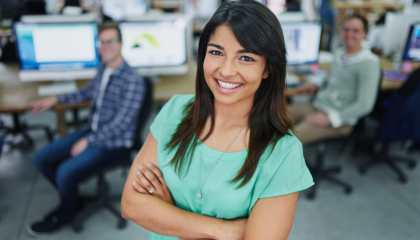 A young person posing in front of the camera. Behind we can see twho people sitting byt the desk in front of computers.