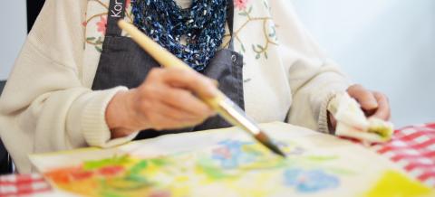 Iäkäs nainen istuu pöydän ääressä ja maalaa siveltimellä väriä kankaalle.
