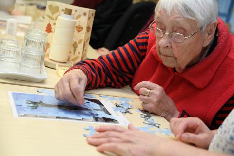 Punapaitainen iäkäs nainen istuu pöydän ääressä tekemässä palapeliä.