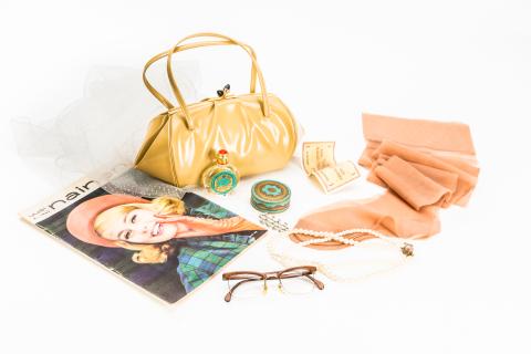 Keltaisen käsilaukun edessä on naistenlehti, silmälasit, sifonkihuivi, helmet ja nailonsukat.