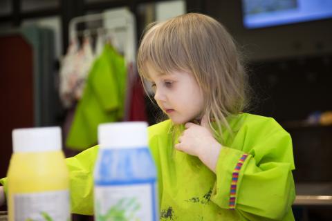 Takaa-alalla lapsi viChild wearing a green apron, and in front of her yellow and blue paintcan. 