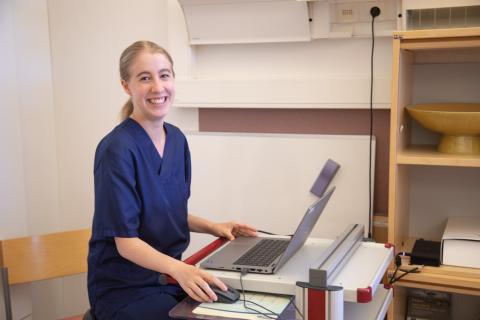 Sairaanhoitaja istuu tietokoneen ääressä ja katsoo kameraan hymyillen.