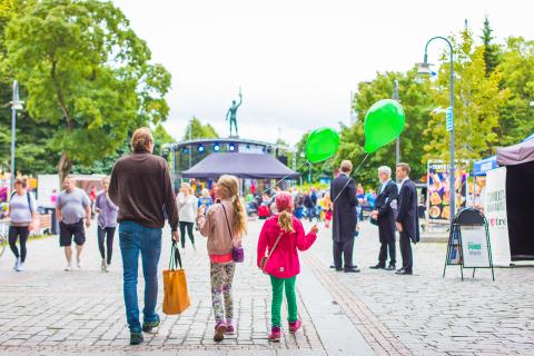 Aikuinen ja kaksi lasta Hämeenpuistossa, lapsilla vihreät ilmapallot kädessä. Taustalla ihmisjoukkoa.