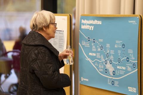 Vanha nainen tutkii PeltsuMultsu kehittyy -julistetta, jossa kuvataan alueen kehityskulkua.