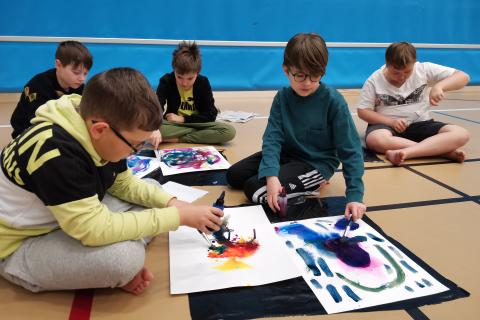 Viisi 6. luokkalaista tekee värikkäitä maalauksia liikuntasalin lattialla. 