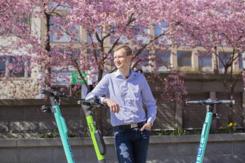 Henkilö seisoo osittain näkyvien sähköpotkulautojen kanssa kukkivien kirsikkapuiden edessä.