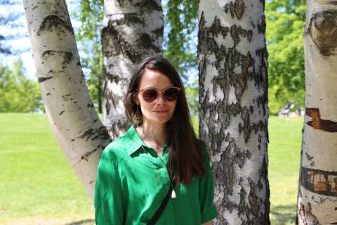 Henkilö vihreässä paidassa ja aurinkolaseissa seisoo puistossa koivujen edessä.