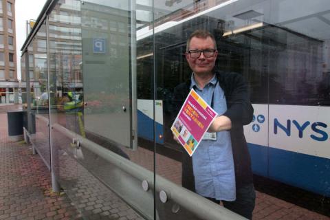 Mikko Ala-Kapee at a bus stop holding a Let&#039;s make Tampere equal together -poster, with a bus in the background.