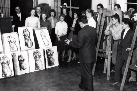 Opettaja seisoo opiskelijoiden edessä. Rivissä on piirrettyjä torsotöitä.