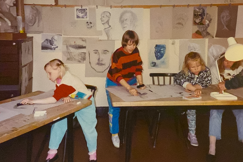 Kolme lasta piirtää pöydän ääressä. Taustalla on kuvataidetöitä ripustettuina seinälle.