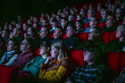 Aikuista yleisöä katsomassa elokuvaa, istuvat punaisilla penkeillä.