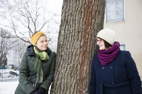 Emmi Nieminen ja Elina Seppänen nojaavat puuhun ja katsovat nauraen toisiaan.