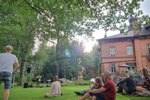 Muusikko esiintyy puistossa kesällä. Kuuntelijat istuvat nurmikolla.