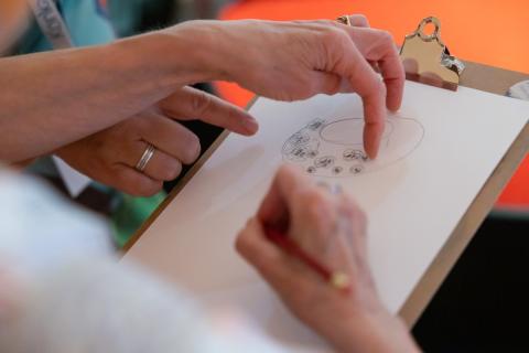 Lähikuvassa kahden naisen kädet piirtämässä samalle paperille hattusuunnitelmaa.