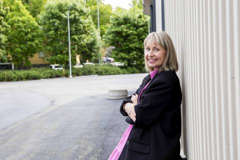 A person leans against a wall and smiles. There are trees in the background.