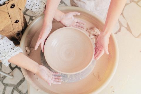 Keskellä kuvaa savesta muotoiltu keskeneräinen astia, jota työstävät kahdet kädet.