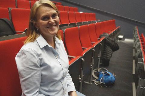 Nainen istuu ja hymyilee auditorion penkillä.