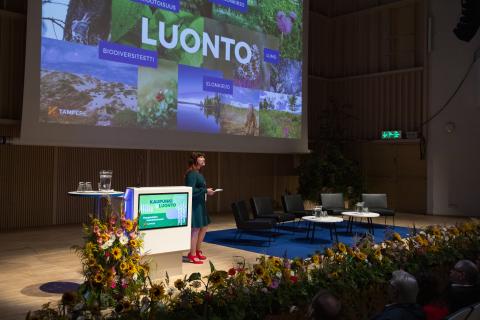 Puhuja isolla lavalla Tampere-talossa taustallaan kuvia sisältävä esitys.