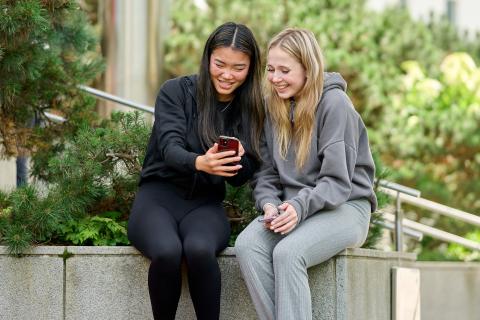 Two girls are sitting outside, laughing and looking at their phones.