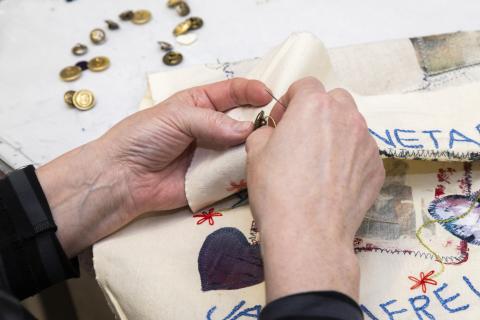 Lähikuva käsistä, jotka ompelevat nappia kirjontatyöhön. Kuvassa näkyy tekstiiliä ja nappeja.