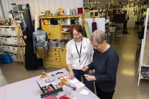 Käsityöpajassa ohjaaja ja pajaan osallistuva katselevat käsityötarvikkeita.