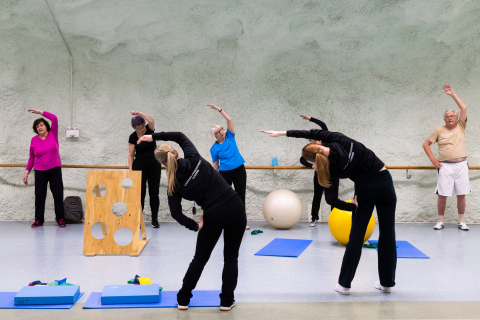 Seniors gymnastics to the rhythm of two instructors in dark clothing.