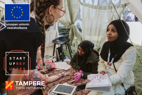 At the event held in the tent, two people are doing henna body art at a table while a third person watches from the sidelines. The table is set with henna supplies and sample designs.