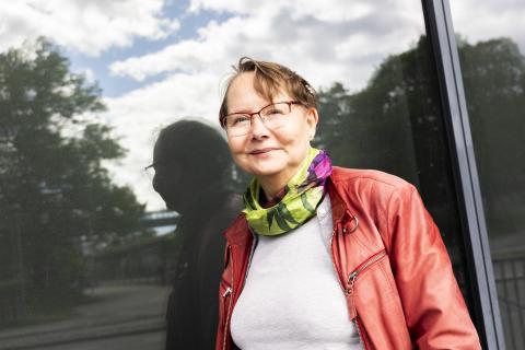 University lecturer Tuula Juvonen, dressed in a red jacket and green scarf, stands outside by the window. Her shadow image is reflected in the window.