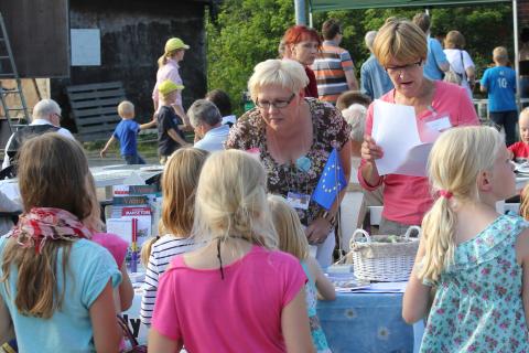 Two city representatives are standing outside at an information table during a summer event. The one with light hair is leaning toward a group of children on the other side of the table.