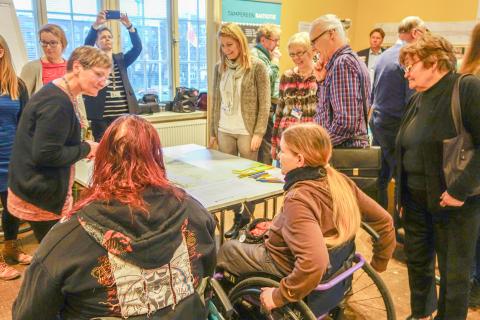 A large group of people is looking at the papers on the table. In the foreground are two people in wheelchairs.
