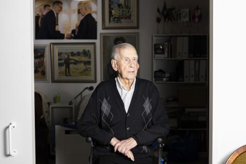 A person sits in front of a wall displaying several paintings and a photograph of the interviewee shaking hands with President Stubb.