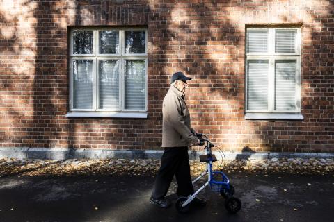 A person wearing a cap walks upright with a rollator. A red-brick wall is visible in the background.