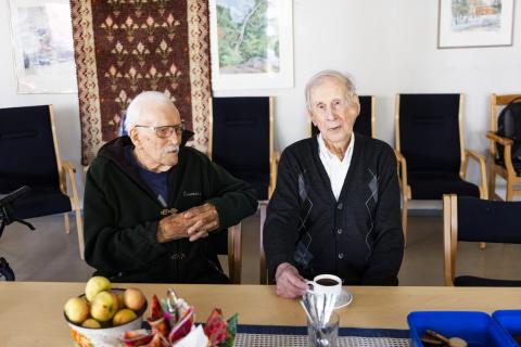 Two people sit at a table. One has a coffee cup in front of them, and there is a bowl of apples on the table.