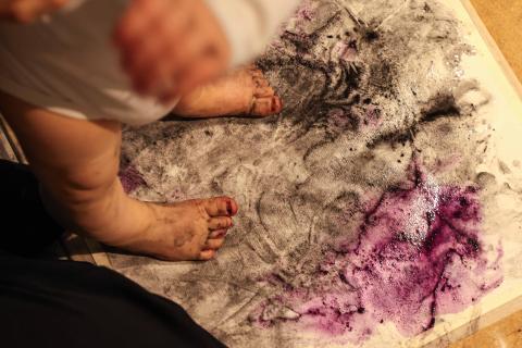 Vauva seisoo värikylpyteoksen päällä lattialla jalat maalissa. Teoksessa käytetty mustana värinä hiiltä ja violettina punajuurta. 